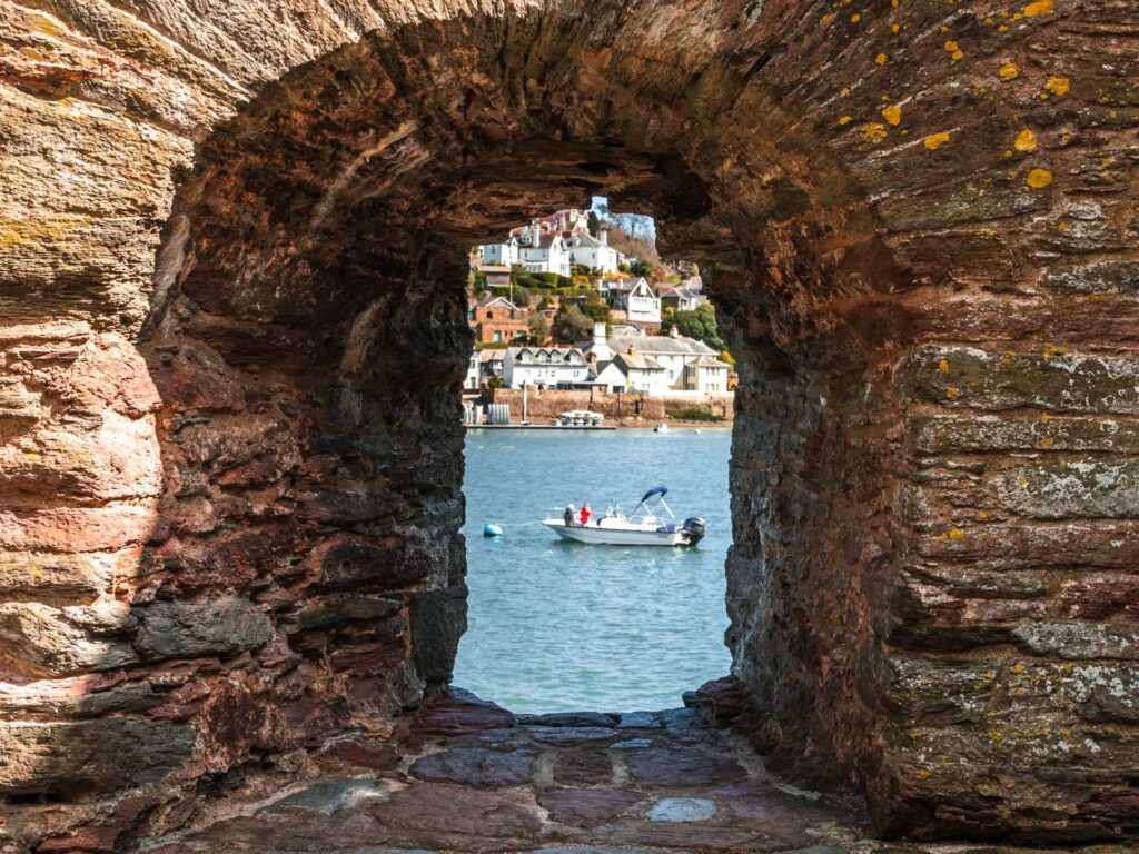 Looking through the brick window hole to the river dart. There is a boat visible through the hole and buildings on the other side of the river.