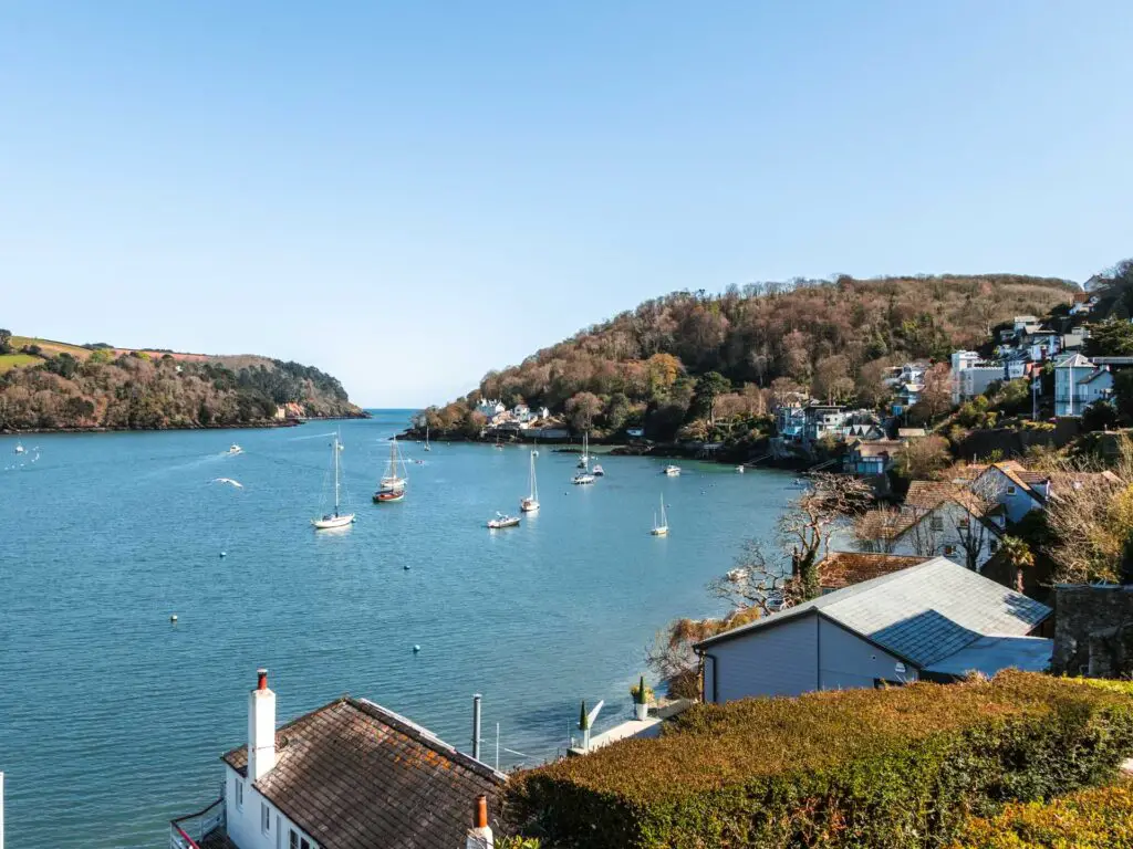 The river dart with boats scattered on it at the start of the walk to Dartmouth Castle. The river is lines with buildings and greenery. The sky is blue.