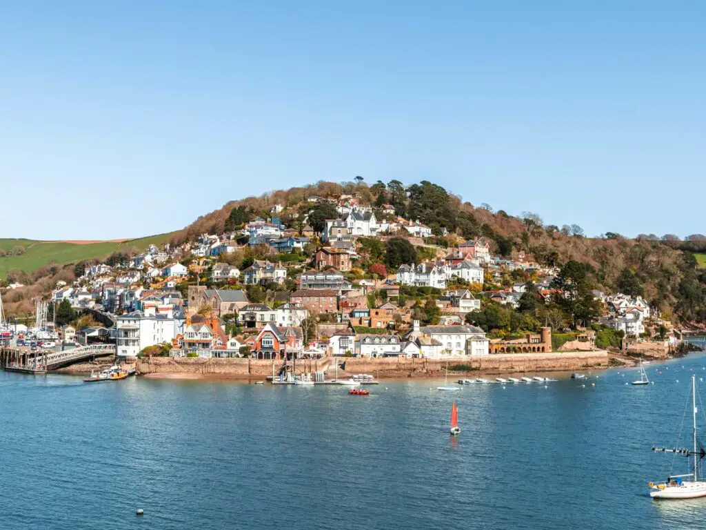 Lots of houses and building on a hill across the blue river dart on the walk to Dartmouth Castle. There are a few boats in the water. The sky is blue.