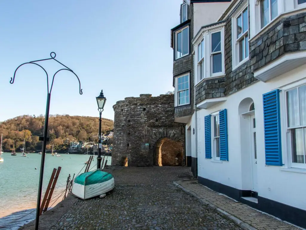 The cobble tree leading to a brick fort. There is a white house on the right with blue window shutters. The river dart is on the left. There is an upturned boat with a turquoise bottom on the path.