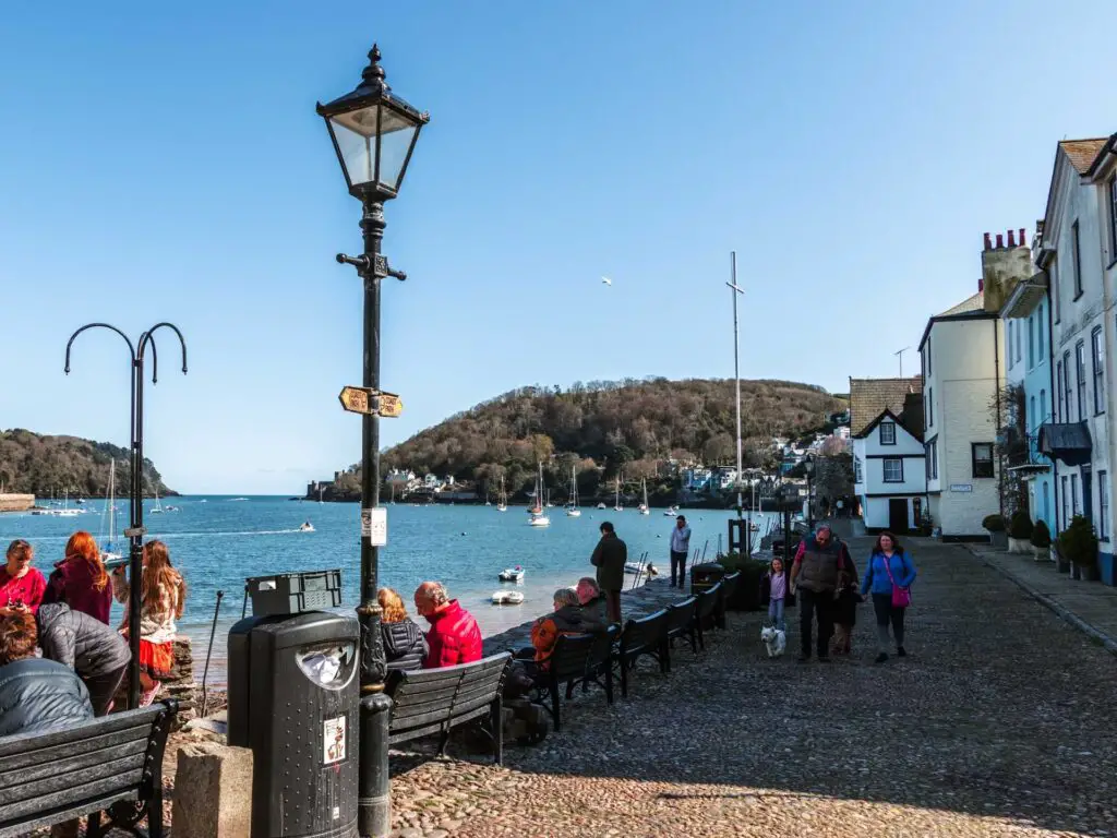 A cobble road with benches lining it looking out to the river Dart. There are people walking on the path and sitting on the bench.