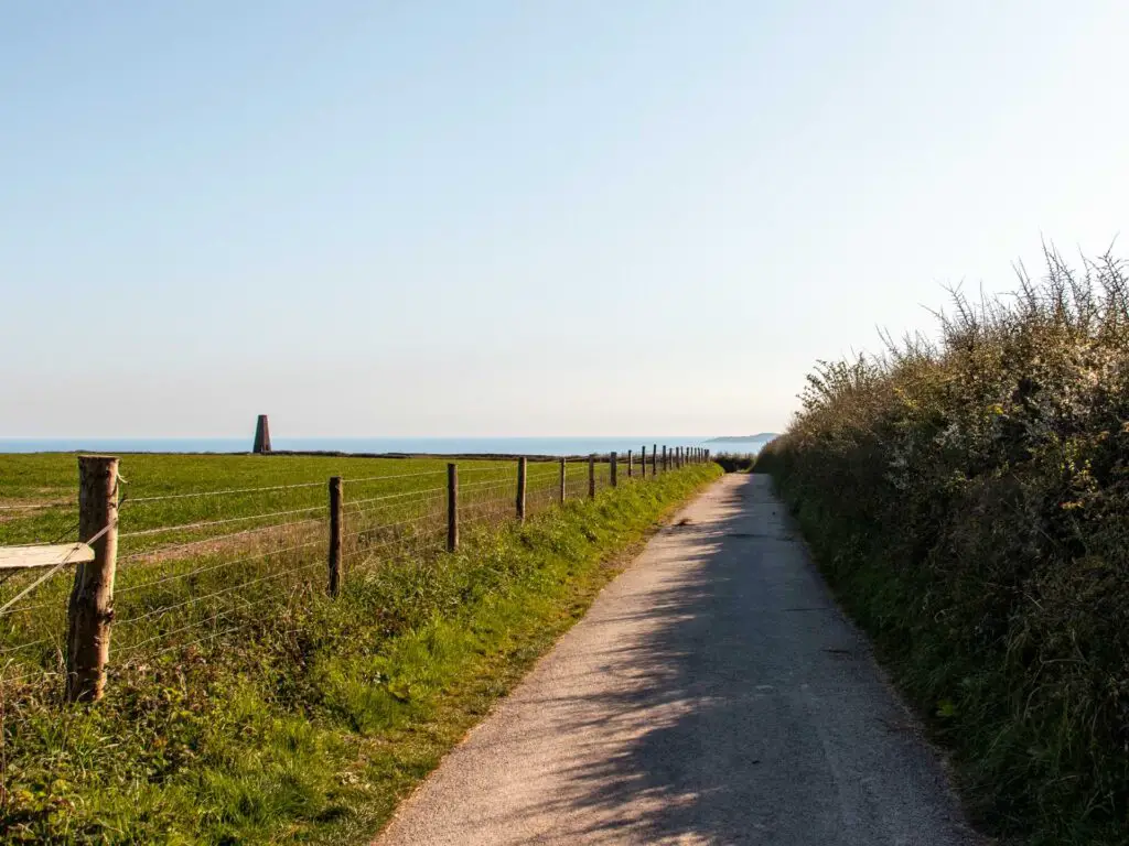 The path leading along the side of a green field with the Daymark tower and blue sea visible in the distance.