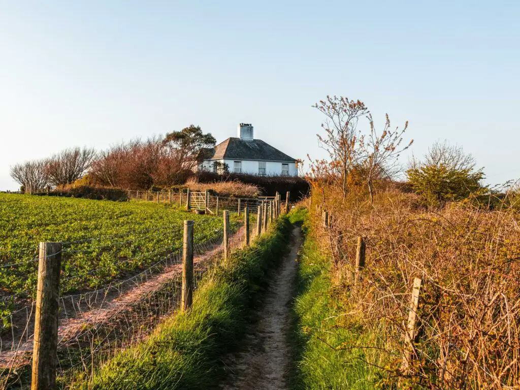 A narrow dirt trail leading to a white house at the end of the Daymark walk near Dartmouth. There is a wire fence on the left side of the trail and shrubbery on the right side.