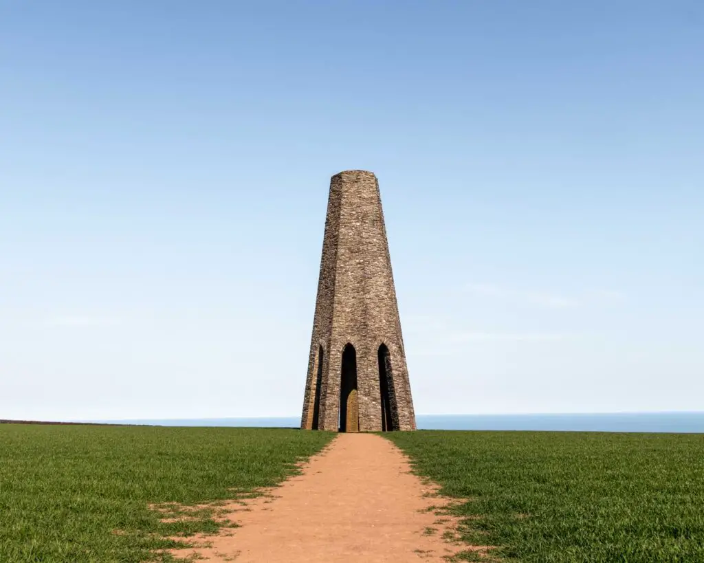 The stone Daymark Tower on the hike near Dartmouth. it is at thew end of a dirt trail and surrounded by green grass. The sky is blue.