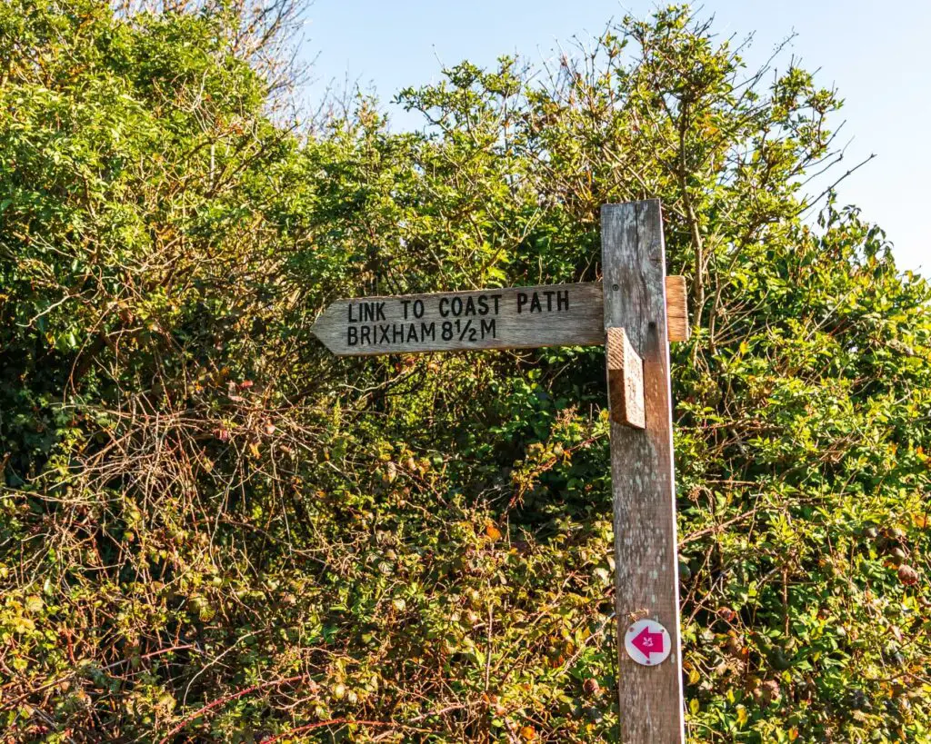 A wooden coast path signpost pointing to Brixham. There is a green bush behind the signpost.