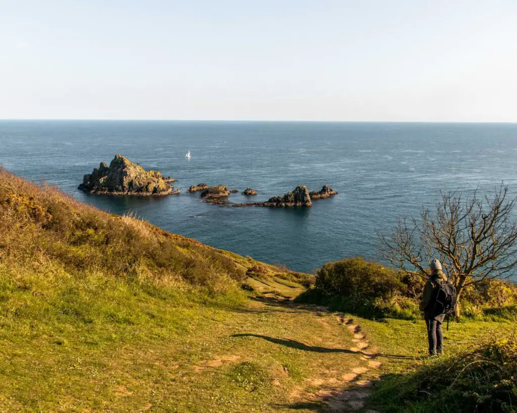 Looking downhill along the green grass to the blue sea. There are a few rocks in the sea. There is a man standing admiring the view.