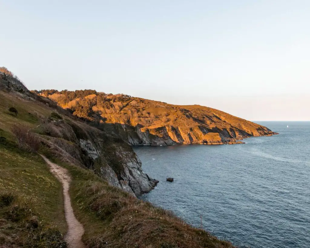 A narrow walking trail running along the side of the rocky cliffs on the hike near Dartmouth. The cliffs are next to the blue sea.