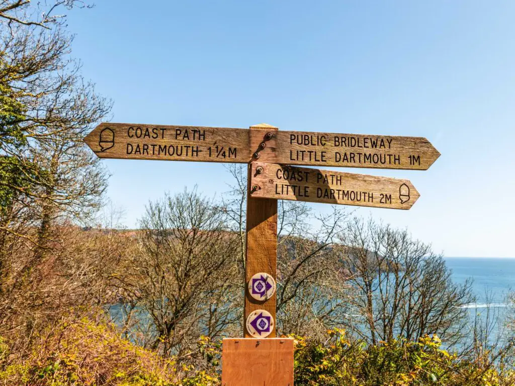 Wooden South West coast path signage pointing to Dartmouth and Little Dartmouth. The sea is visible in the background. 