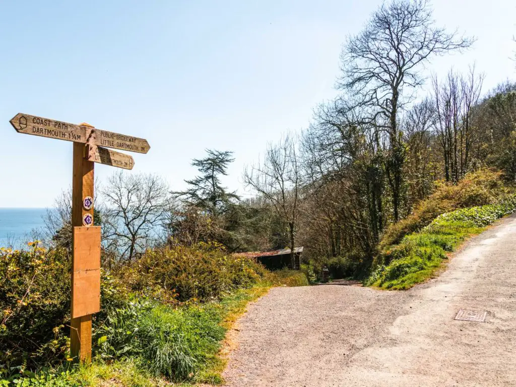 A wooden South West coast path signpost pointing to two trails. There is green grass on the side of the trails and the blue sea in the background.