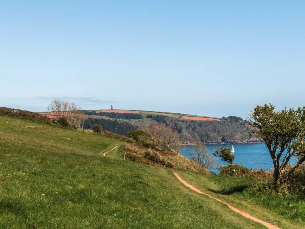 A narrow coastal path trail running through the hilly green grass with the blue sea on the right and the Daymark visible on the hill in the distance. 