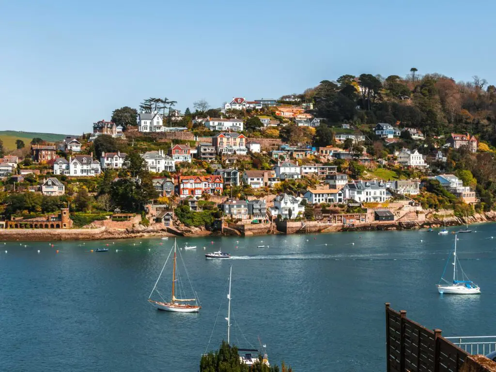 Looking across the blue River Dart to the houses on the hill on the other side on the walk to Little Dartmouth. There are a couple of sail boats on the river.