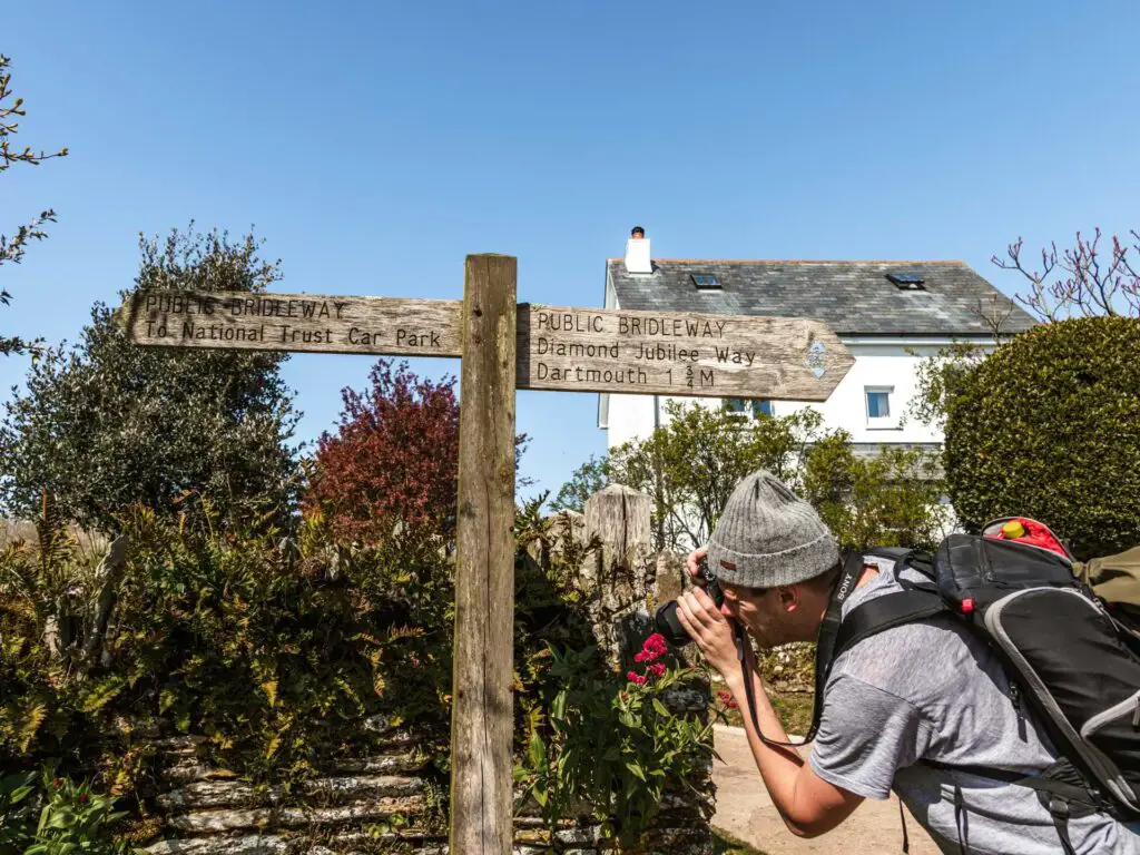 Wooden South West coast path signpost with a white home in the background. Ollie is standing next to the signpost photographing the flowers.