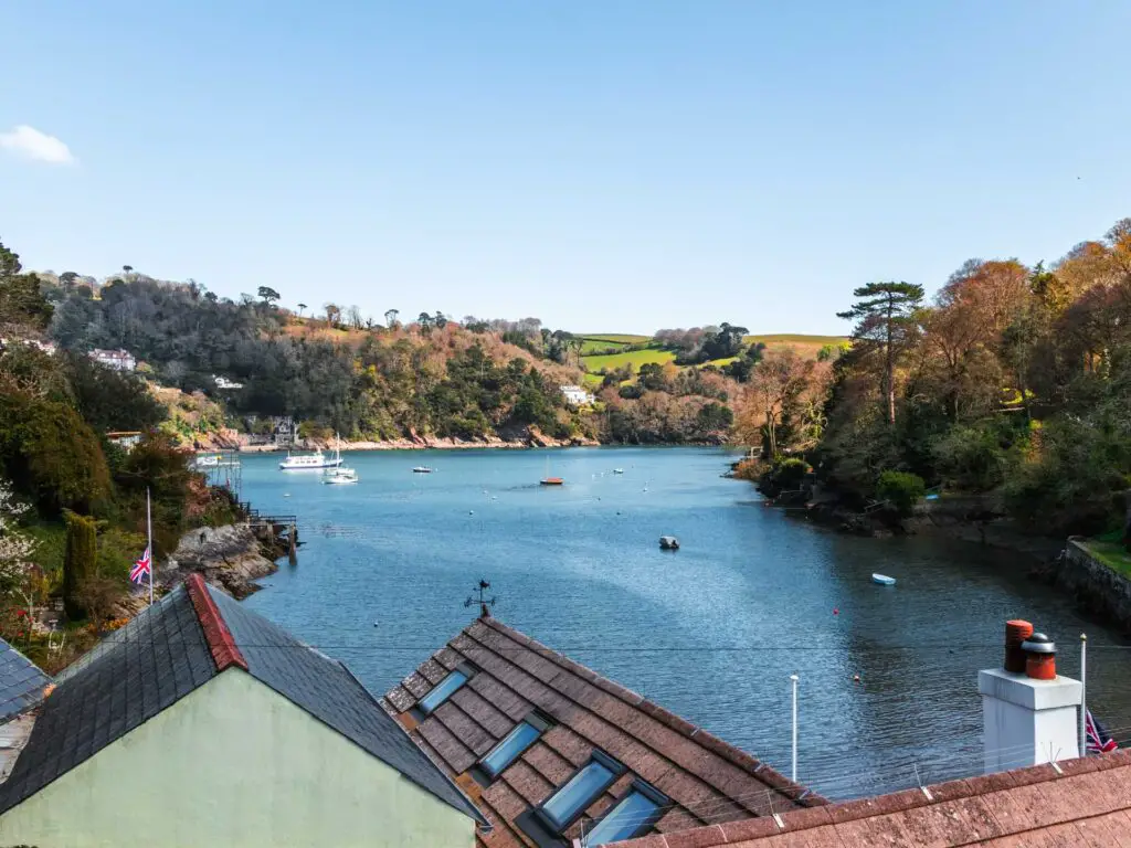 Looking over the rooftops to the blue River Dart surrounded by green land and trees. There are a few boats on the water.