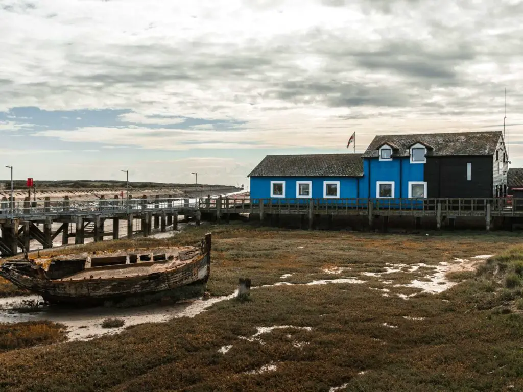 A derelict boat and a blue and black building on the walk into Rye Harbour.