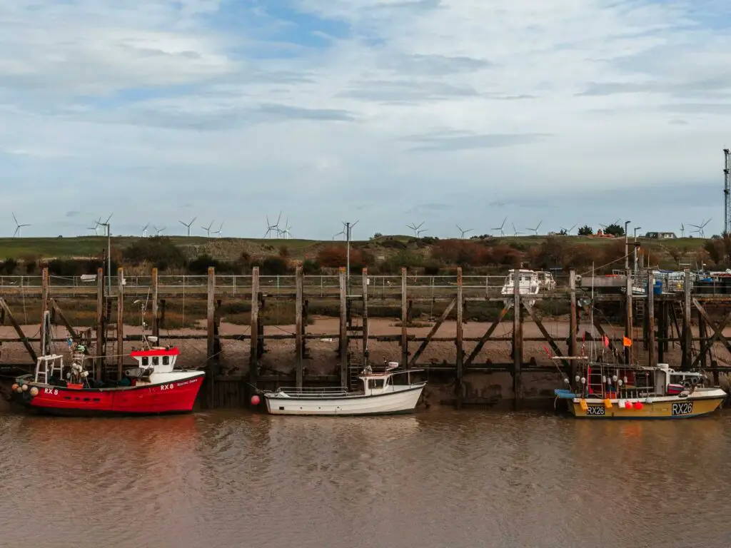 Boats floating on the brown coloured river at Rye Harbour. There is a wind farm visible in the distance.