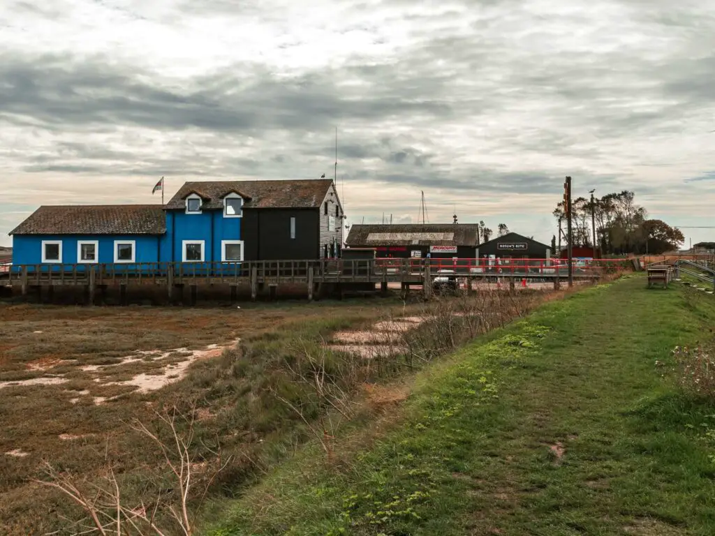 A green grass walking trail with the blue and black building ahead to the left at Rye harbour.