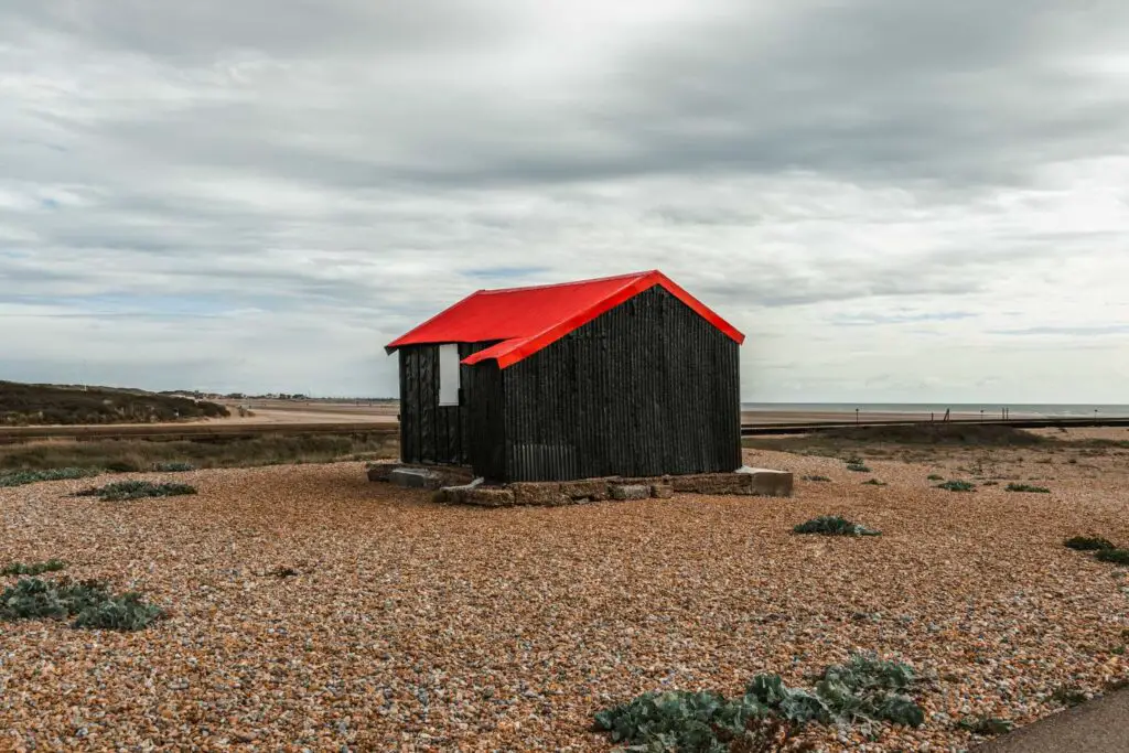 The black wood shed with a red roof on the Rye Harbour walk. It is sitting on a pebble ground.