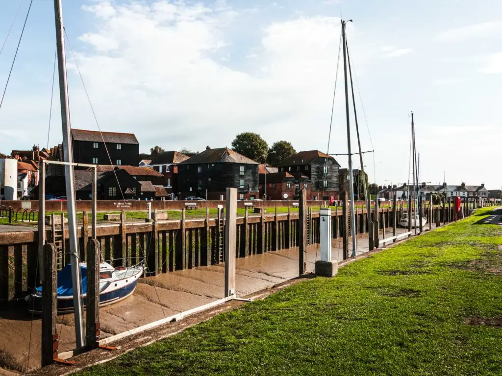 The river bed with a sail boat resting on it. There are black buildings on one side and a grass bank on the other.