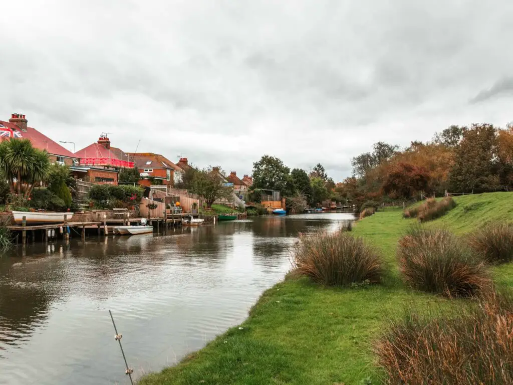 A river with a grass back on the right side and houses on the left side on the walk back from Camber Castle to Rye.