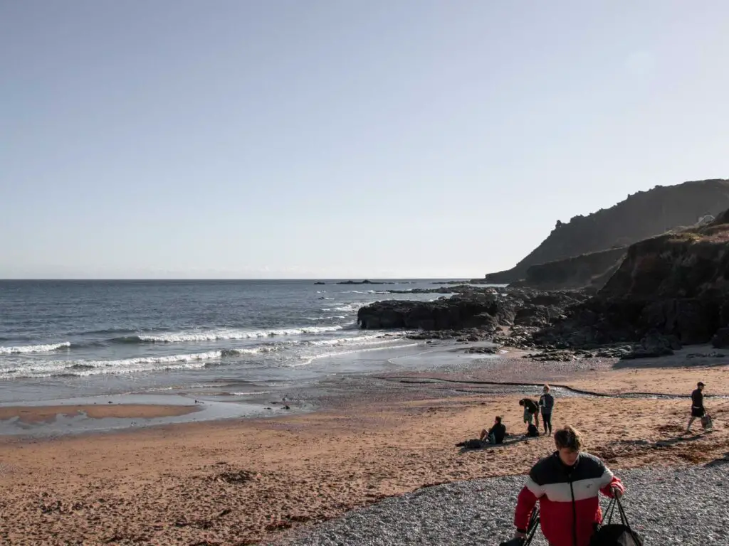 A sandy beach with cliffs to the right side. There are a few people walking on the beach. The sea is blue.