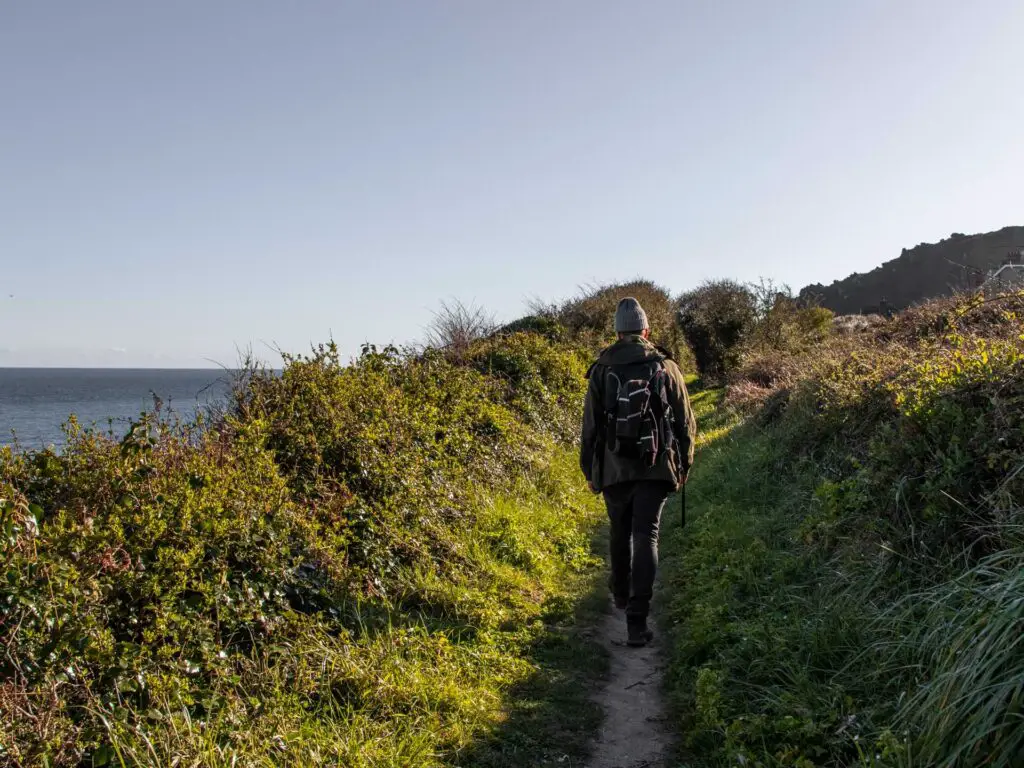 A man walking along the dirt trail of the south west coast path. There is green grass and bushes on the sides of the trail, with the blue sea on the left.