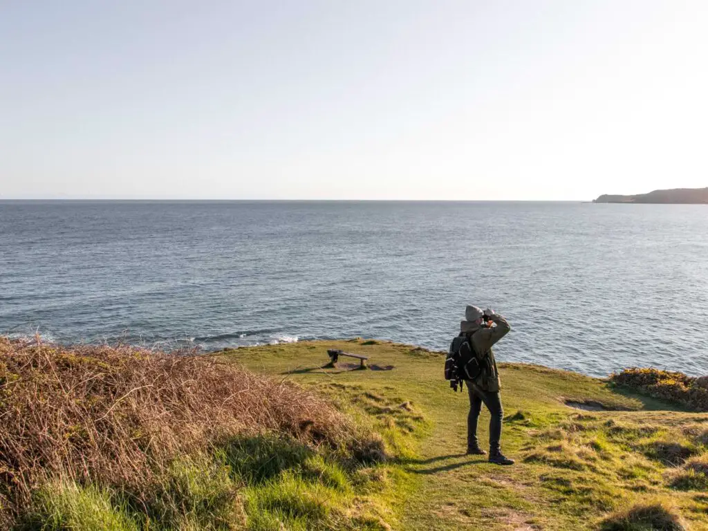 A man standing on the green grass taking a photo of something off frame. The blue sea is in the background.