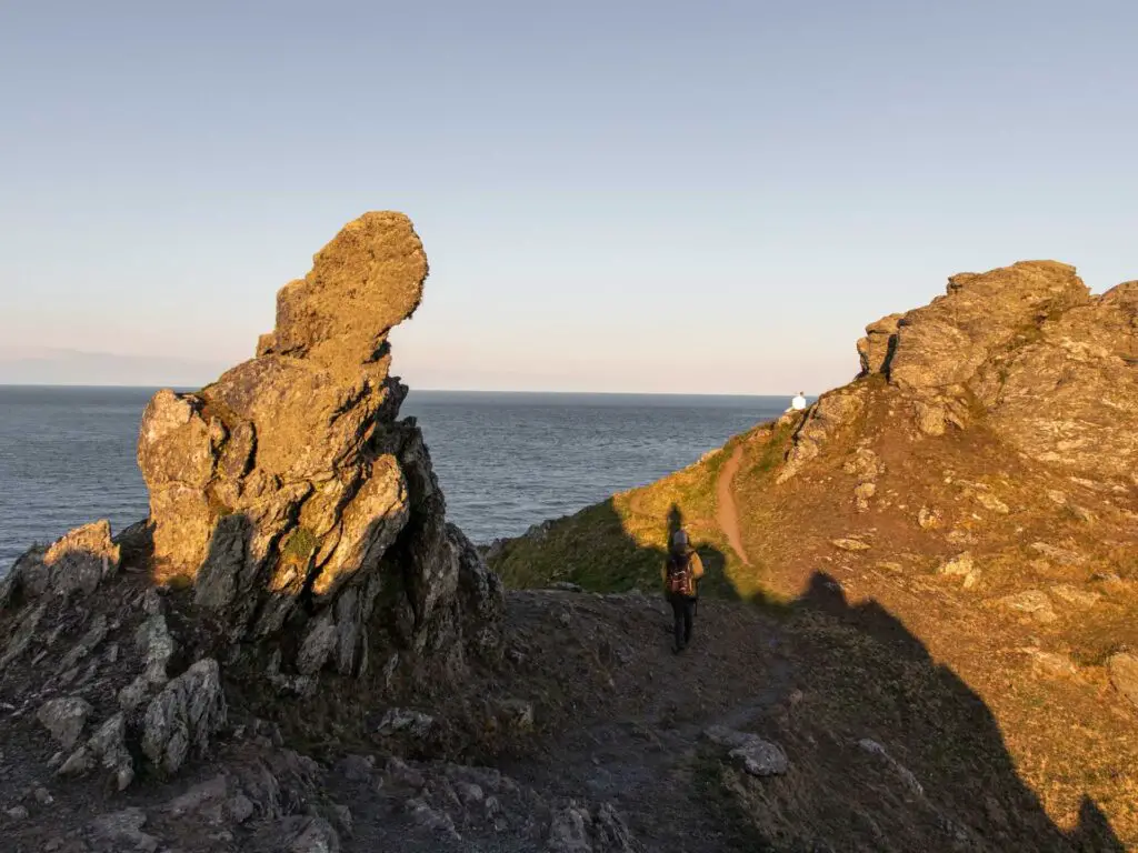 A man walking on the ridge with jagged rocks behind and ahead of him on the start point lighthouse walk. The blue sea is in the background.