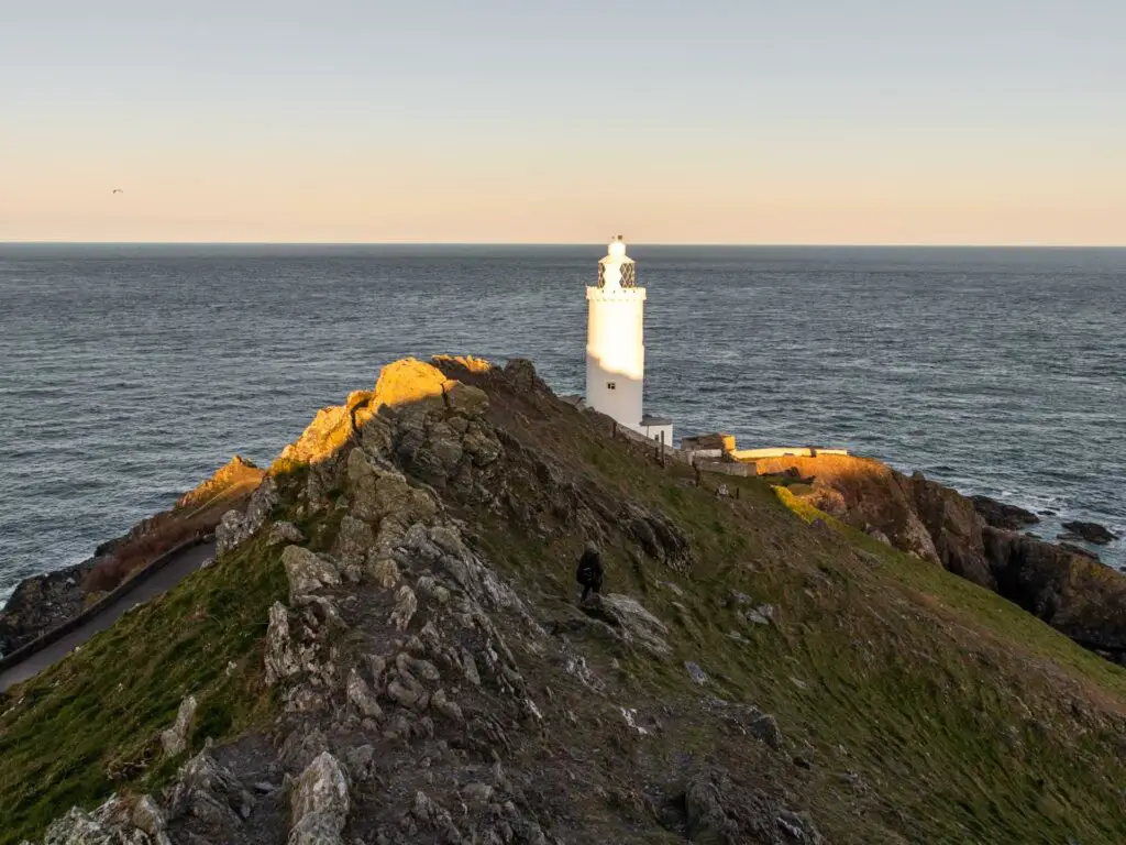 A man walking across the side of a rocky hill towards Start point lighthouse with the sea in the background.