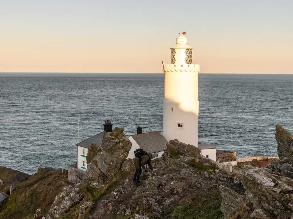 A man walking across the rocks with the white start point lighthouse in front. The blue sea is the backdrop.