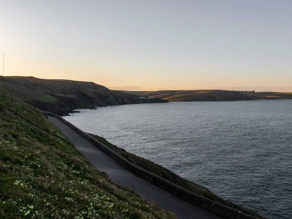 The road running along the bottom of the green grass hill with the sea below it to the right. There is a view along the coastline as the sun is setting.