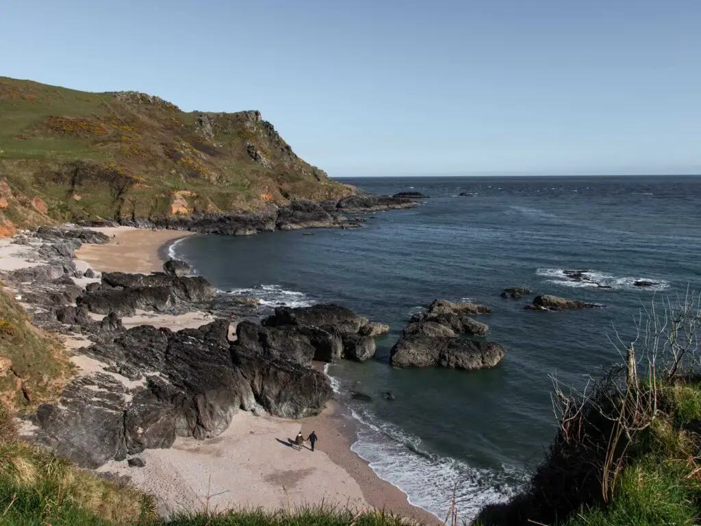 A sandy beach Brocken up by big black rocks with a hill in the background on the Start Point lighthouse walk. The sea is blue. There re two people walking on the beach.