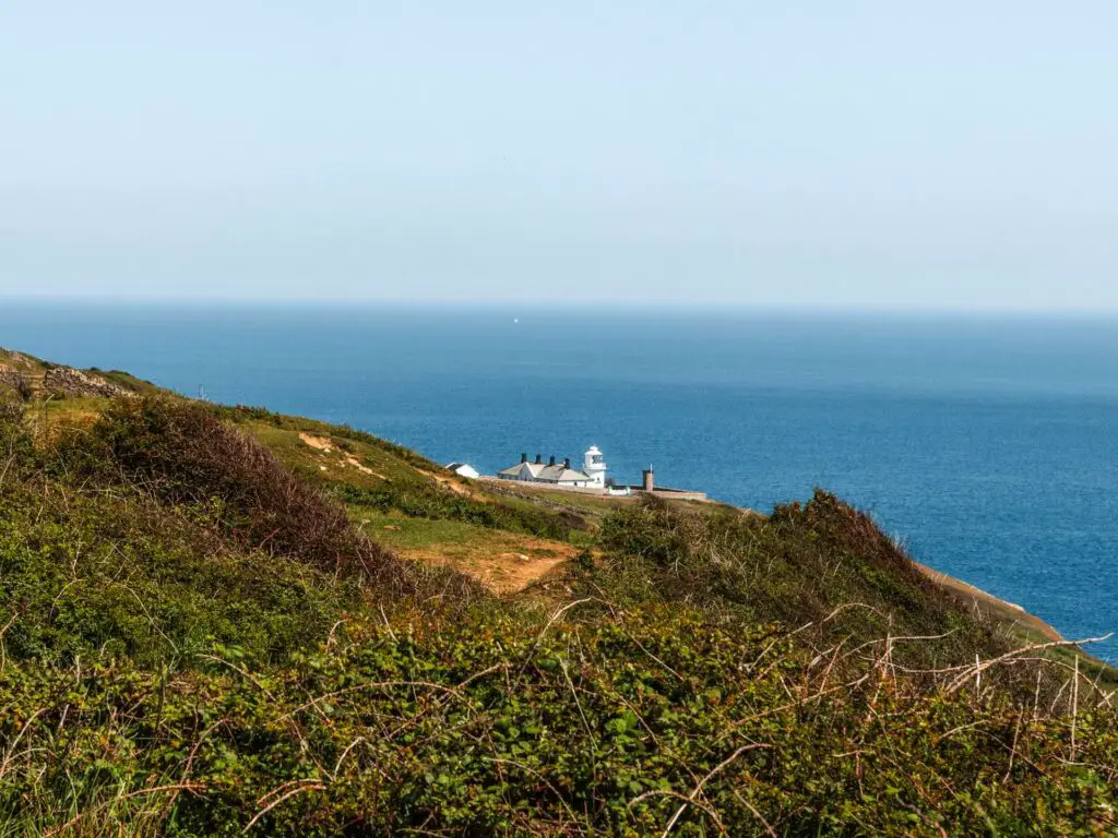 A big green hill with lots of bushes and Anvil Point Lighthouse half visible on the other side. The blue water of the English Channel is behind it.