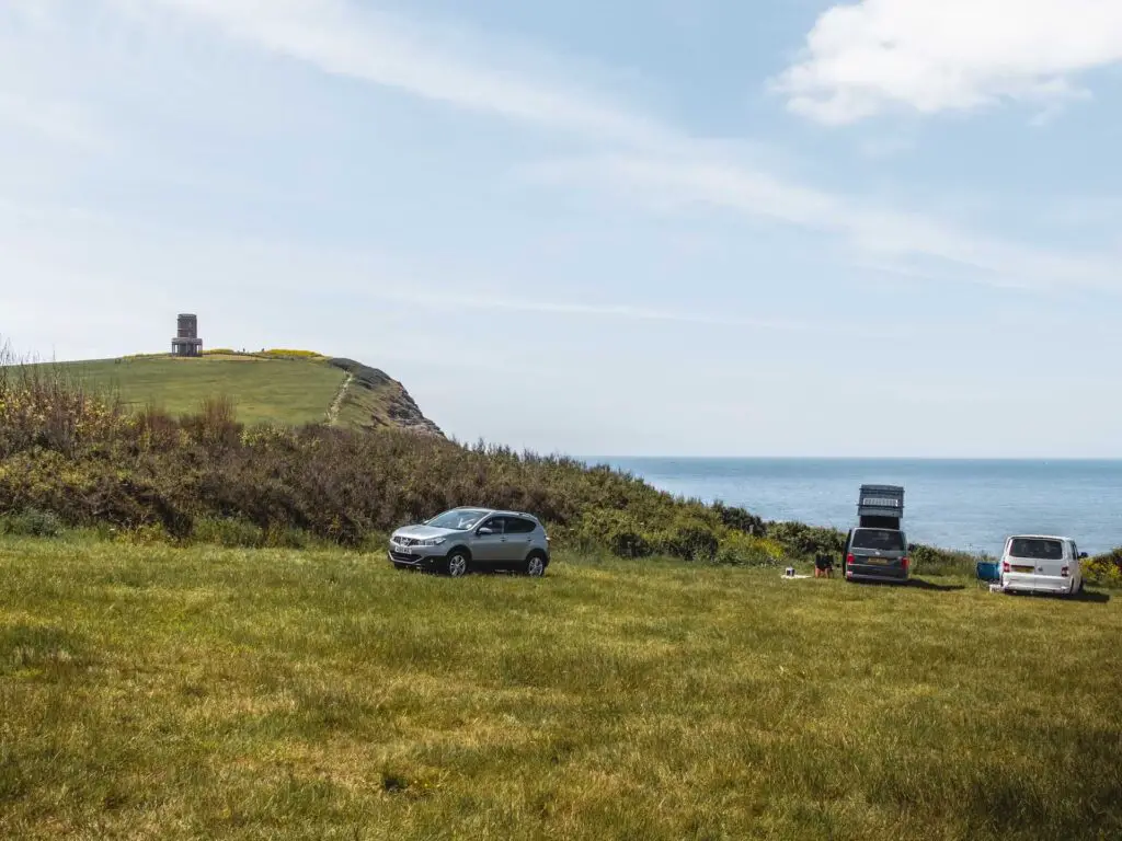 A green grass filed car park with a view to the sea and a hill with clavell tower on top. There are a few cars parked.