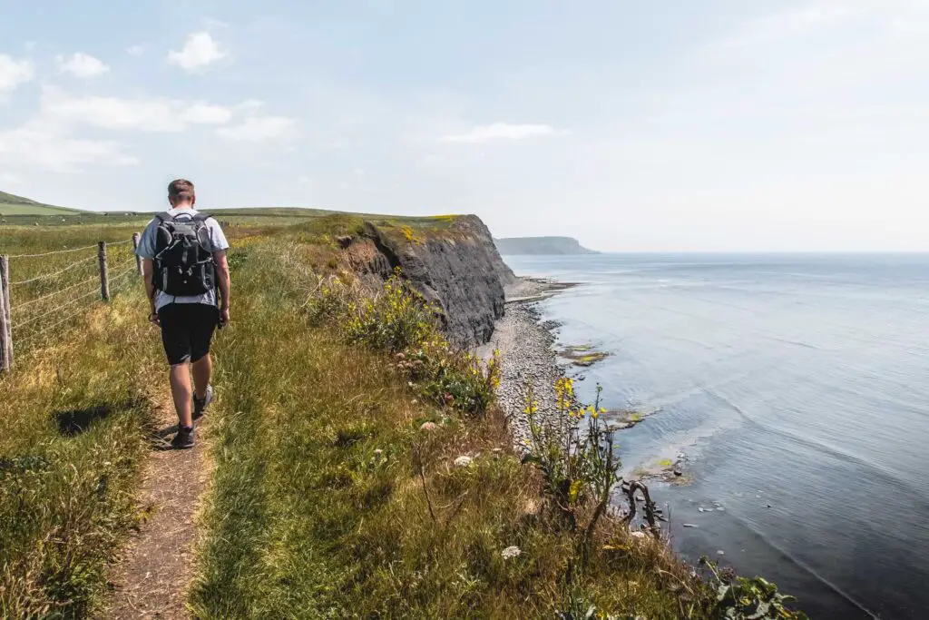 A man walking on a small dirt trail on the edge of the cliff on the coastal walk from Kimmeridge. The cliff leads down to the sea.