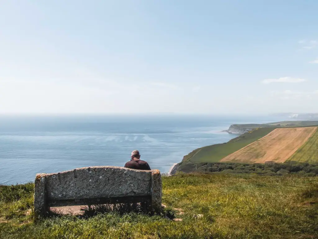 A stone bench on the hill top looking downhill along the coastline in Dorset. There is a man sitting on the bench.