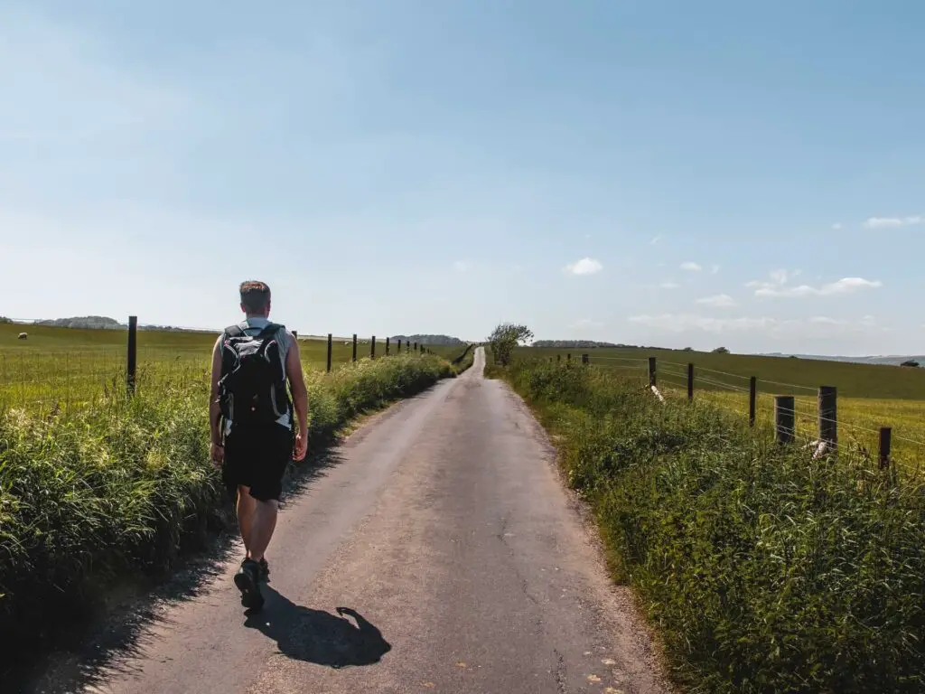 A man walking along a long road lined with fences and tall grass with a field on both sides.