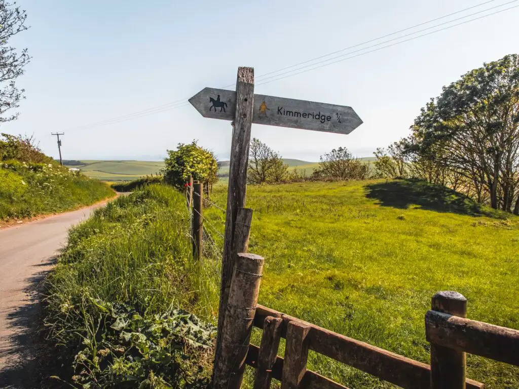 A wooden signpost on the side of the road, pointing to kimmeridge with a picture of a walking man.  