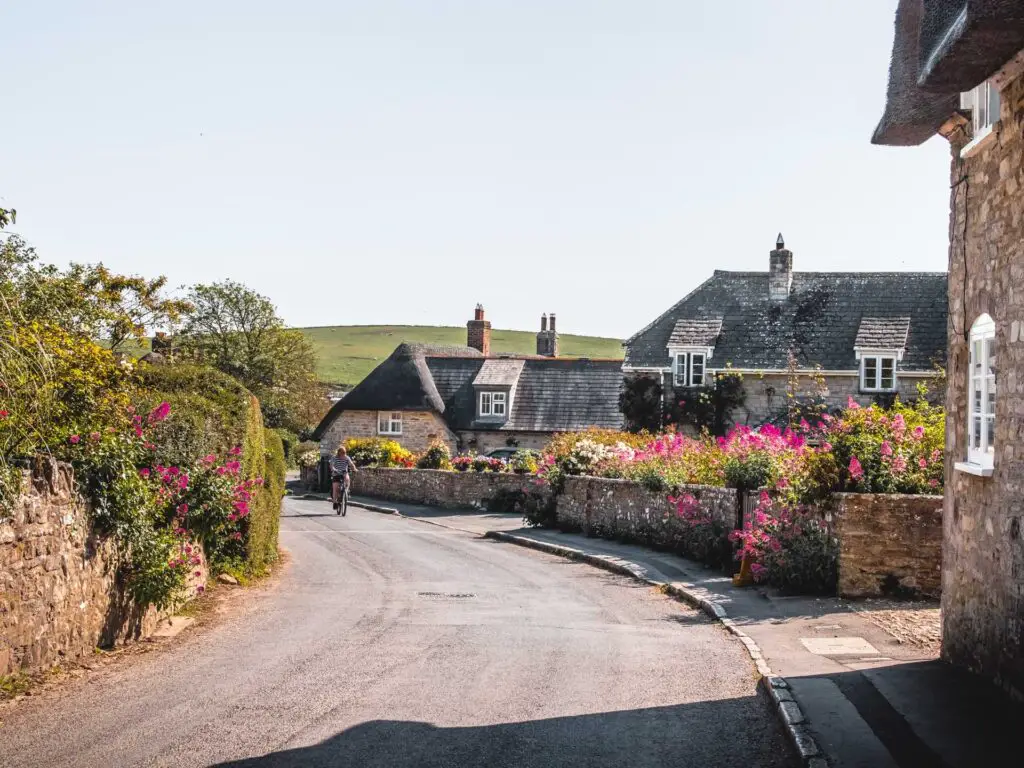 A road curbing through Kimmeridge Village. The road is lined with brick and flint houses, with walls and pretty flowers. There is a person cycling on the road.