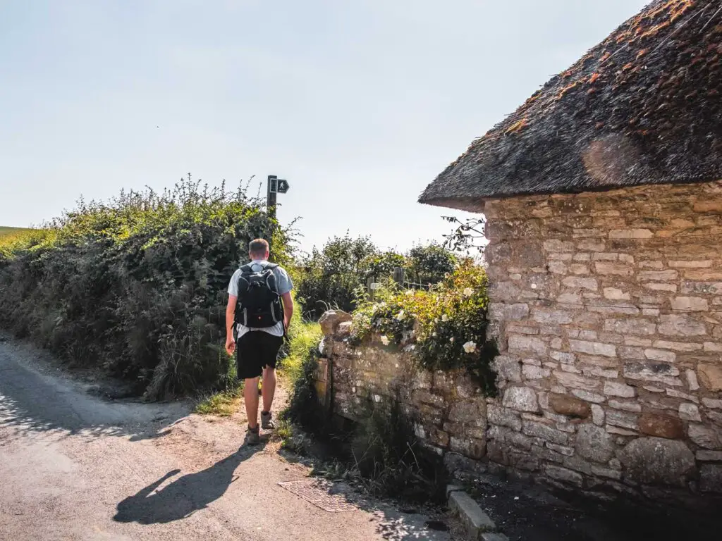 An opening between a hedge and stone building. A man is walking through the opening where there is a signpost.