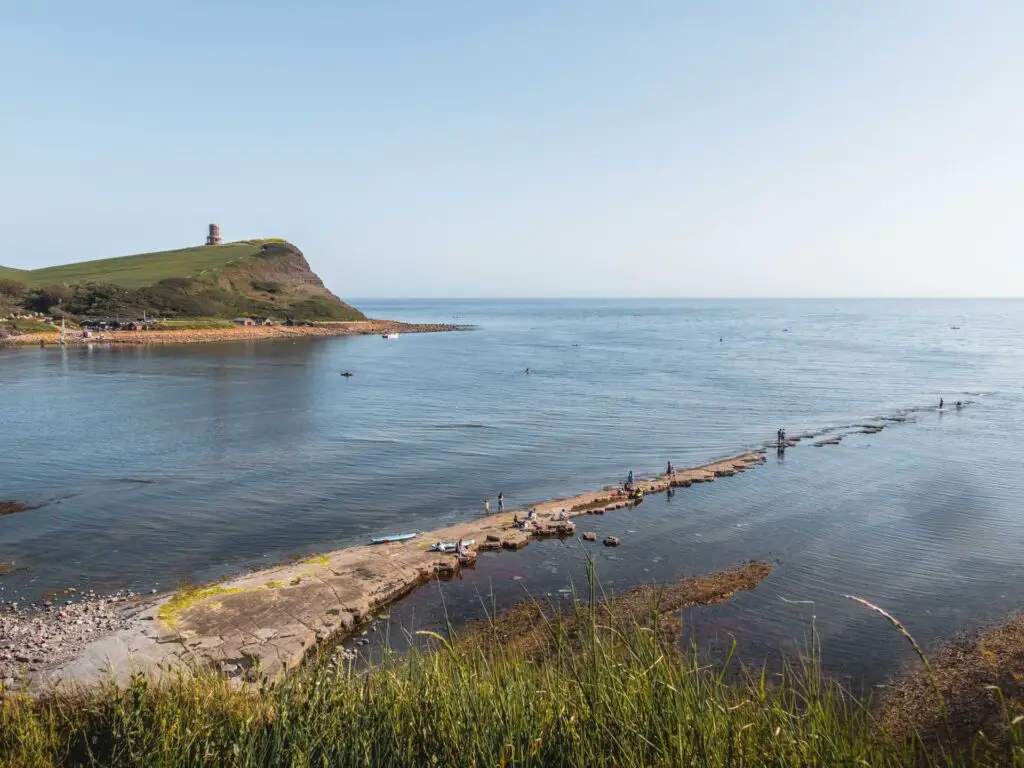 Looking across the sea of Kimmeridge bay and people walking along rock slabs and a hill with Clavell tower on top in the distance. 