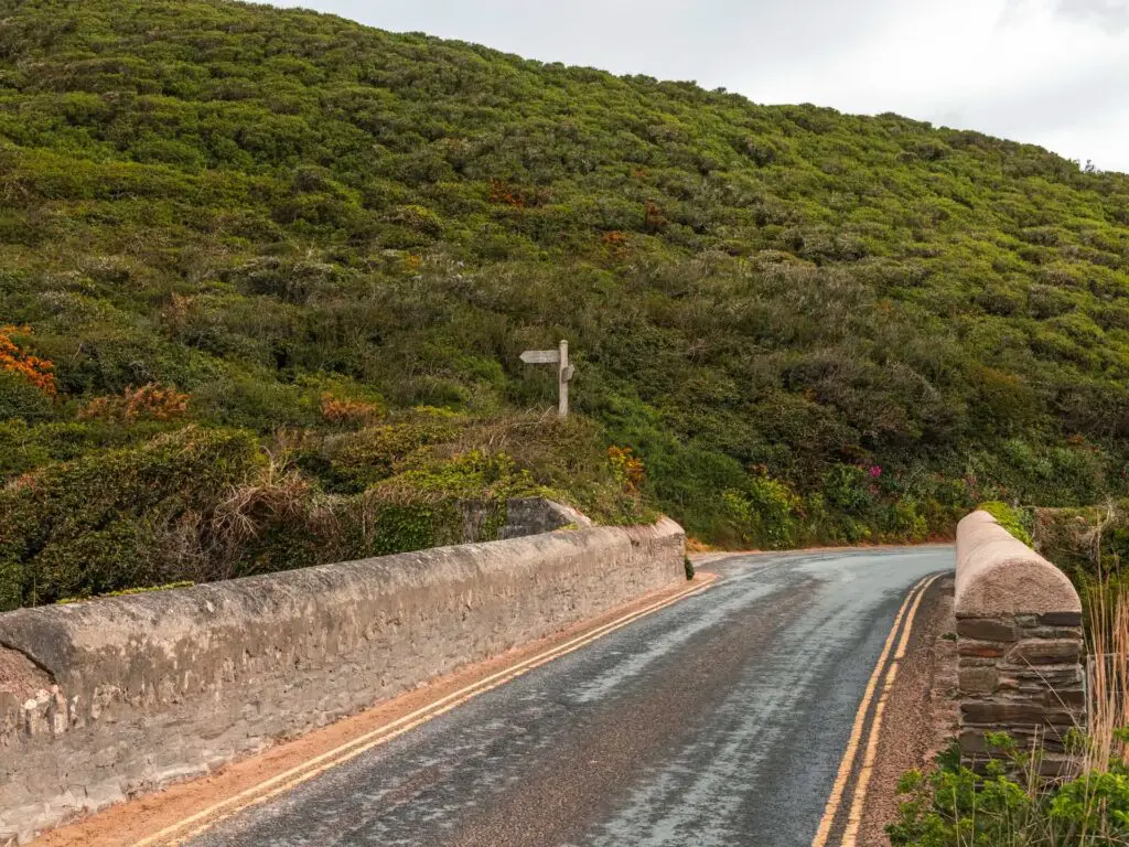 A road over a bridge with a green bushy hill on the other side.