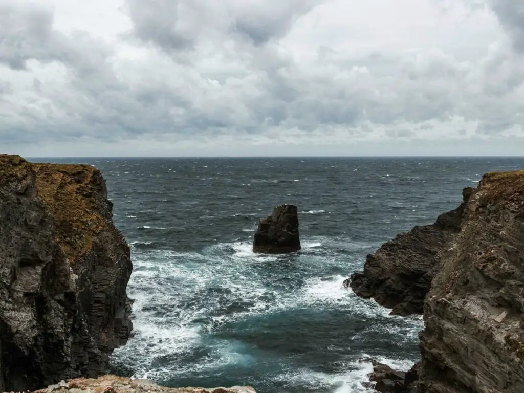 Black rock cliffs on the left and right with the blue sea in-between. There is a small black rock in the middle, sticking out of the sea.