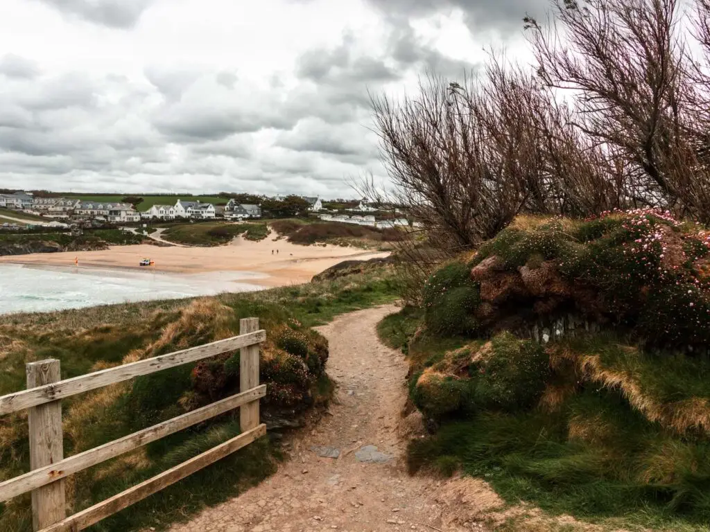 A dirt trail winding through a wooden fence on the left, and grass and moss covered wall on the right. Treyarnon beach is visible in the distance. 