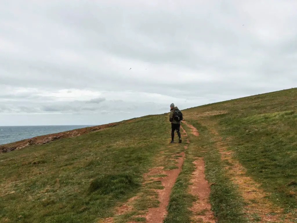 A man walking across the dirt trail on a grass hill.