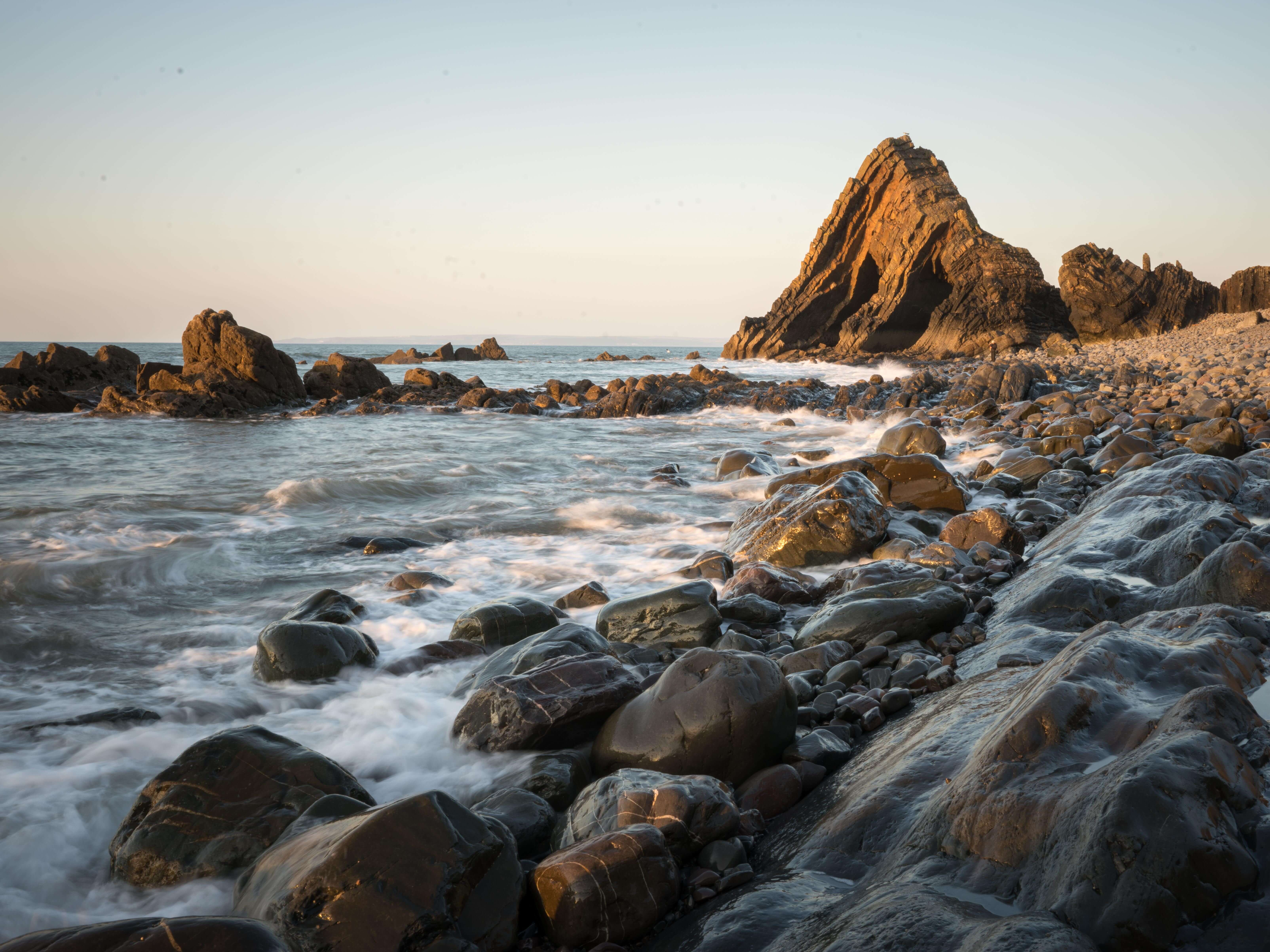 Black rocks with the water rushing over them and Blackchurch Rock in the distance on the Mouthmill Beach walk