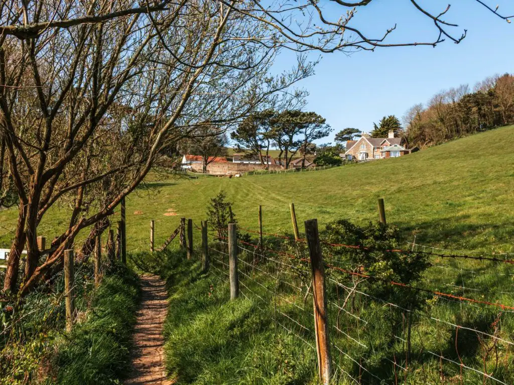A small dirt trail running through the grass and lines with a fence. There is a field on the other side of the fence and a house in the distance. 
