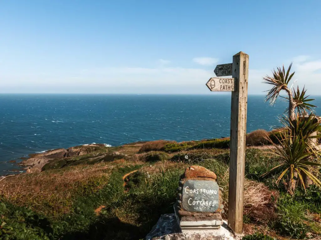 Wooden coast path sign post infront of the green hill which leads to the blue sea.