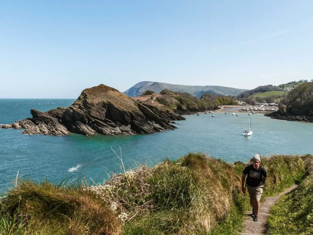 A view of the peninsula of watermouth bay on the walk from Ilfracombe. There are a few boats in the water. There is a man walking along the trail.