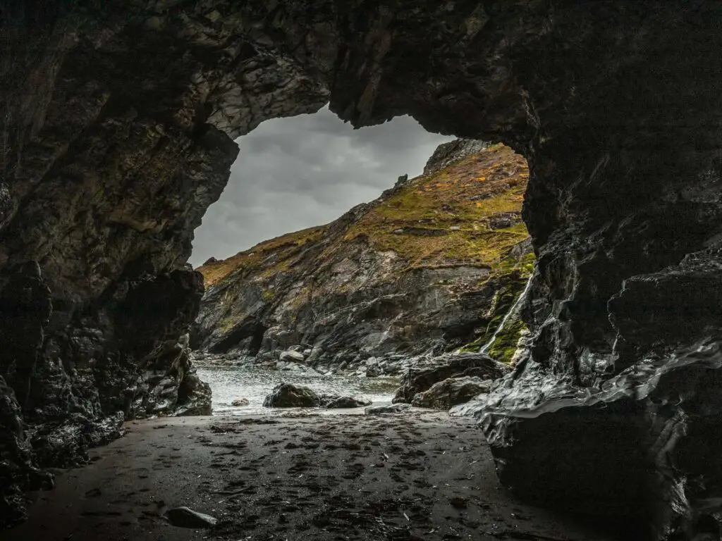 Looking through the opening of Merlins Cave. The cave is dark, with a sandy ground. Looking outside to the rugged cliff where is meets the sea. It is an overcast day.