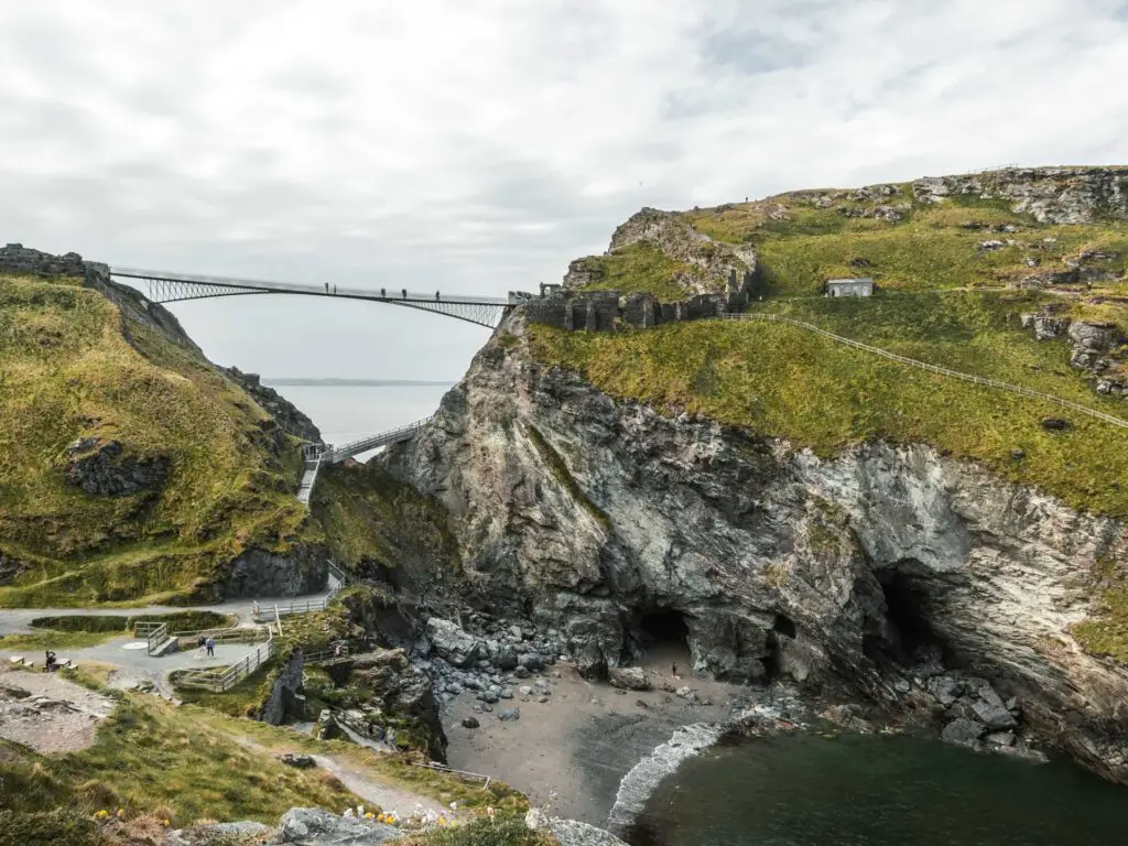 A small beach cove below Tintagel island and people walking across the bridge from the island to to hill on the mainland.